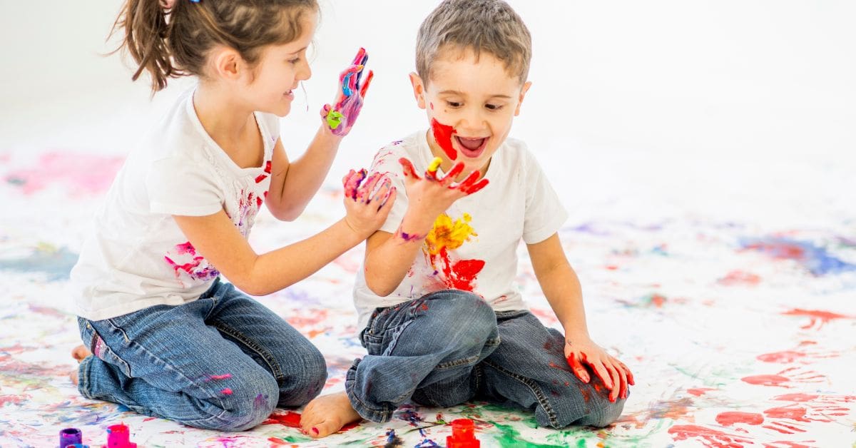 two children in white t-shirts and jeans, playing and smeared in colours. No doubt there is a caregiver around
