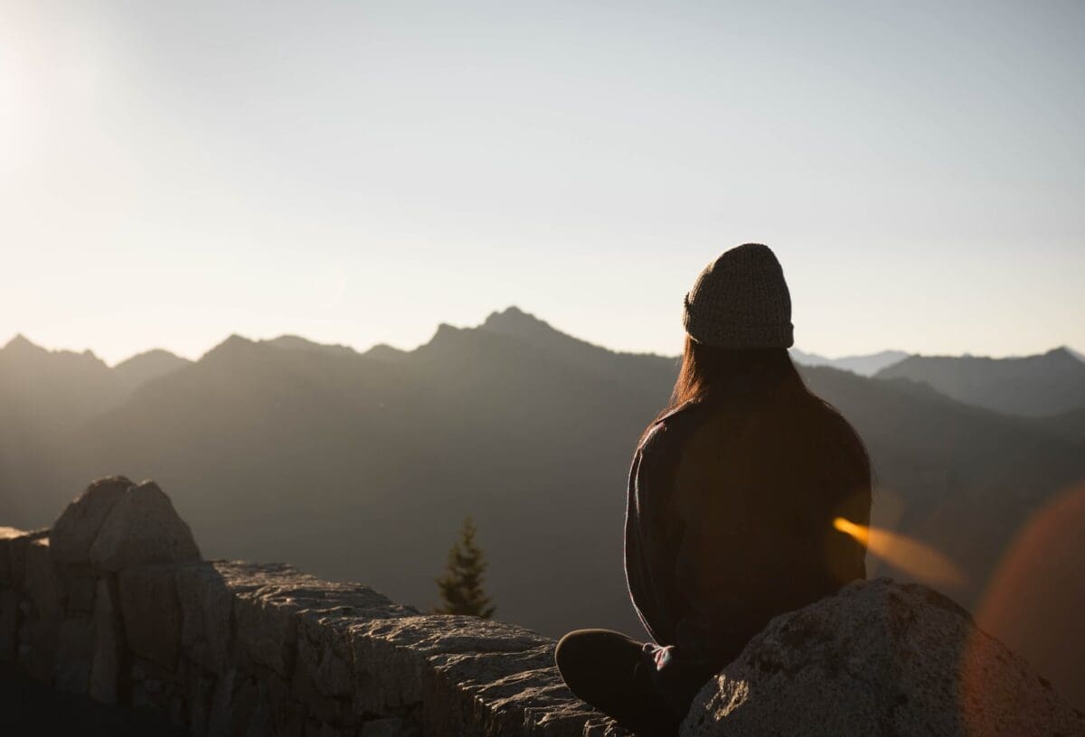 person wearing knit cap facing mountain in a sage like pose
